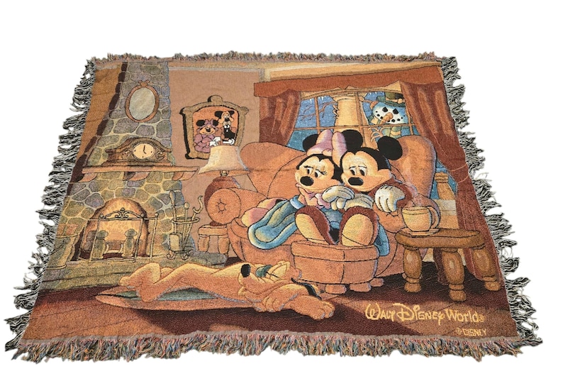 May include: A woven tapestry throw blanket featuring a scene of Mickey and Minnie Mouse sitting on a sofa by a stone fireplace with Pluto sleeping on the floor. The design includes a window with a snowman outside and the text Walt Disney World.