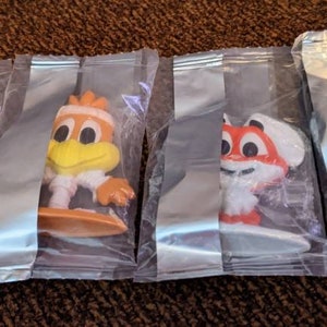 May include: Four small plastic figurines of cartoon characters on surfboards. The characters are a yellow bird, a red fox, a white bird, and a white bear. The figurines are packaged in clear plastic bags.