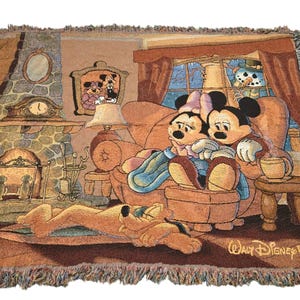 May include: A woven throw blanket featuring a scene of Mickey and Minnie Mouse sitting on a couch by a fireplace, with Pluto sleeping on the floor. The design includes a window with a snowman outside, a lamp, and the text Walt Disney World.