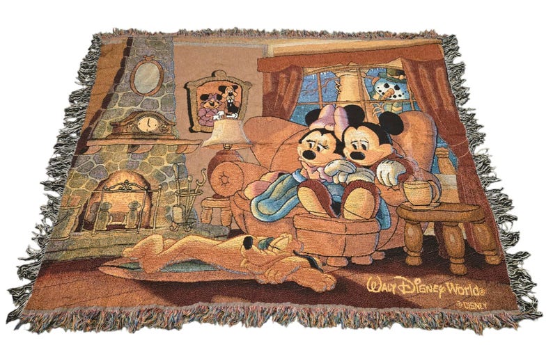 May include: A woven throw blanket featuring a scene of Mickey and Minnie Mouse sitting on a couch by a fireplace, with Pluto sleeping on the floor. The design includes a window with a snowman outside, a lamp, and the text Walt Disney World.