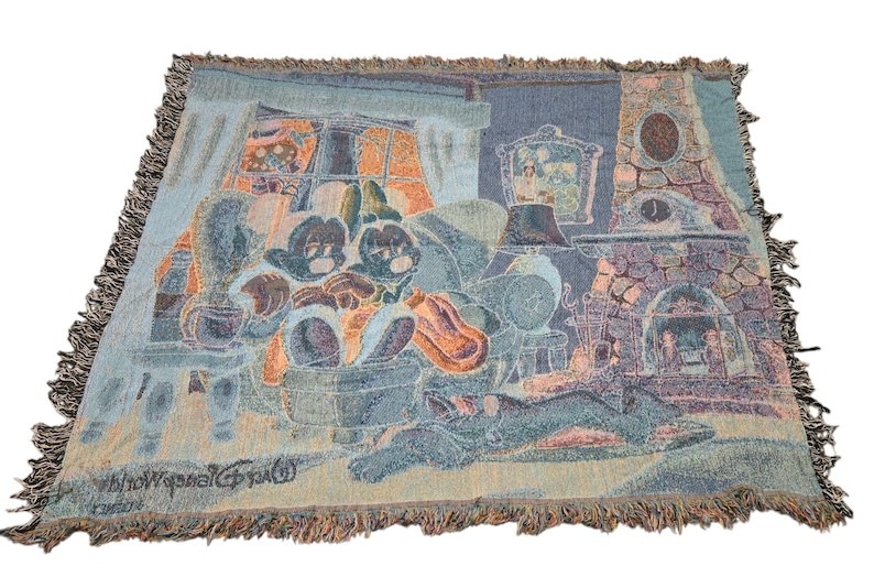 May include: A woven throw blanket featuring a scene of two cartoon mice sitting in armchairs by a stone fireplace. The design includes a window with orange light, a framed picture on the wall, and a dog sleeping on the floor. The text Walt Disney World is visible in the bottom left corner, and the blanket has a fringed edge.