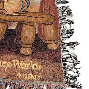 May include: A woven tapestry throw blanket featuring a scene with a wooden table, a teacup, and curtains. The text "Disney Worlds" and "&copy; DISNEY" is visible in gold thread at the bottom. The blanket has a decorative fringe edge with multi-colored threads.