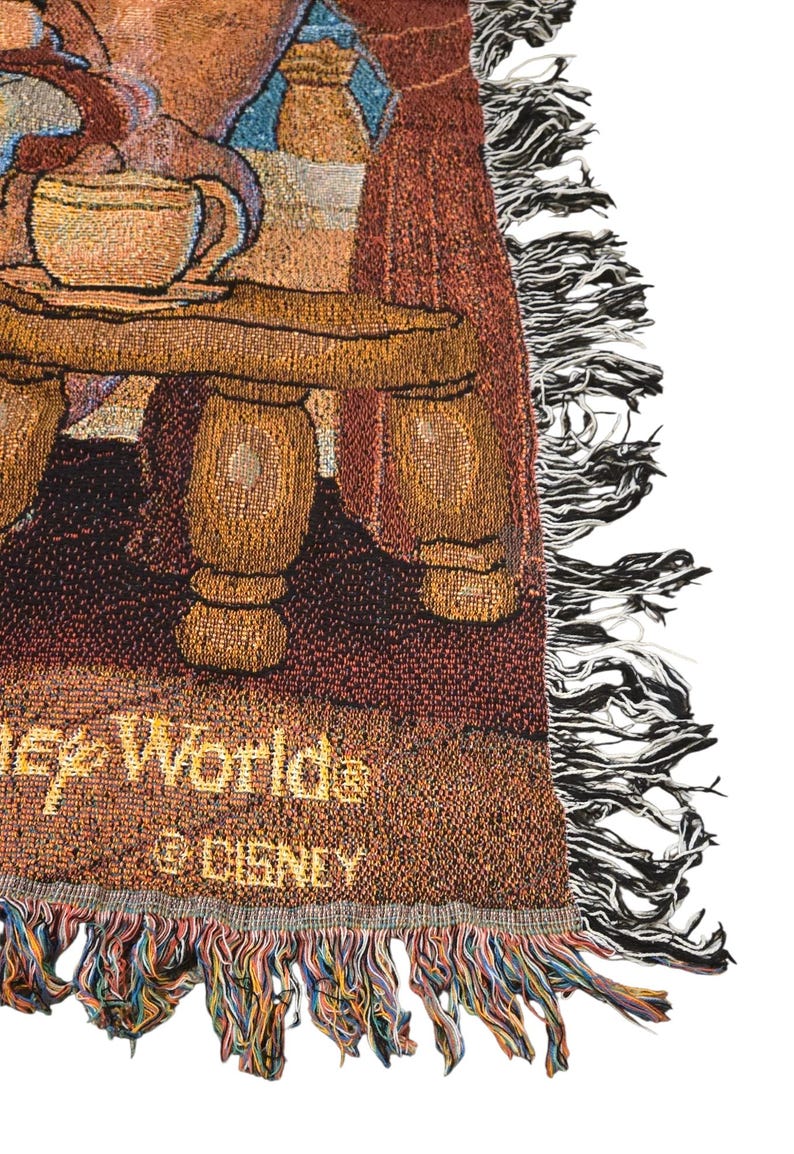 May include: A woven tapestry throw blanket featuring a scene with a wooden table, a teacup, and curtains. The text "Disney Worlds" and "&copy; DISNEY" is visible in gold thread at the bottom. The blanket has a decorative fringe edge with multi-colored threads.