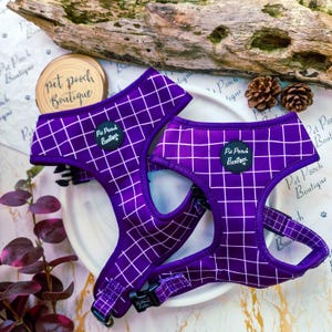 Op de afbeelding: Twee paarse hondentuigjes met een wit ruitpatroon. Elk tuigje heeft een zwart label met de tekst "Pet Pooch Boutique". De tuigjes worden gepresenteerd op een wit bord, met een houten stam en dennenappels op de achtergrond.