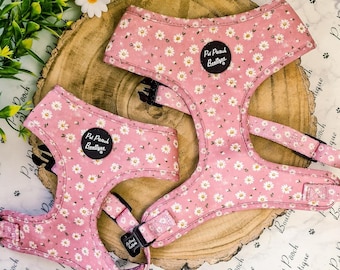 May include: Two pink dog harnesses with a daisy floral pattern. Each harness has black buckles and a black circular patch with the text "Pet Pooch Boutique". The harnesses are displayed on a wooden slice.