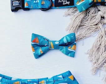 May include: A turquoise blue bow tie with a sailboat pattern. The bow tie is made of fabric and has a small, white, embroidered bird on the right side.