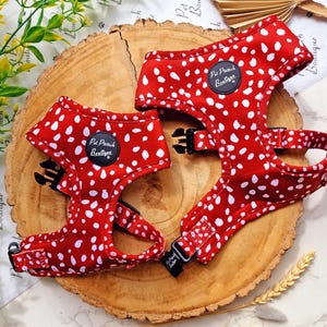 May include: Two red dog harnesses with white polka dots. Each harness has black buckles and a circular patch with the text "Pet Pooch Boutique". The harnesses are displayed on a wooden surface.
