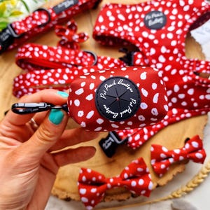May include: Red and white polka dot dog harness, lead, poop bag holder, and bow tie. The poop bag holder has a black circle with the text "Pit Pouch Boutique".