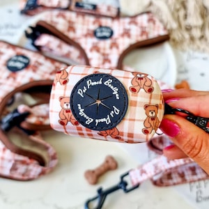 Op de afbeelding: Een dispenser voor hondenpoepzakjes met een bruin en wit teddybeerpatroon. De dispenser heeft een zwarte cirkelvormige opening met de tekst "Pet Pooch Boutique". Een bijpassende riem en tuigje zijn ook zichtbaar.