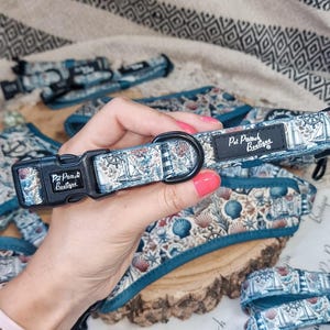 Può includere: Un set collare e imbracatura per cani a tema nautico. Il collare presenta una fibbia nera e un design in tessuto con barche a vela, conchiglie e carte nautiche. Il marchio "Pet Pouch Boutique" è visibile.