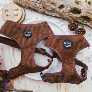 Op de afbeelding: Twee bruine corduroy hondentuigjes met verstelbare bandjes en zwarte gespen. Elk tuigje heeft een cirkelvormige patch met de tekst "Pet Pooch Boutique". De tuigjes worden gepresenteerd op een wit bord met houten accenten.