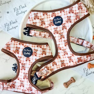 May include: A pet harness featuring a light peach and white plaid pattern with repeating brown teddy bear illustrations. The harness has dark brown trim, black plastic buckles, and a circular navy blue patch with the text Pet Pooch Boutique.