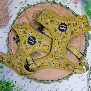May include: Two mustard-yellow dog harnesses with a giraffe pattern. The harnesses have black buckles and the text "Pet Pooch Boutique" on a black patch. The harnesses are placed on a wooden slice with greenery.