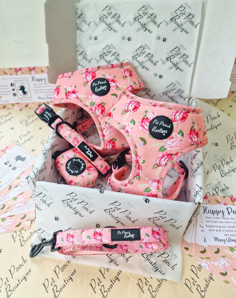 May include: A pink floral dog harness and leash set with a matching poop bag holder. The set is packaged in a box with tissue paper and a card that says "Happy Pooch".