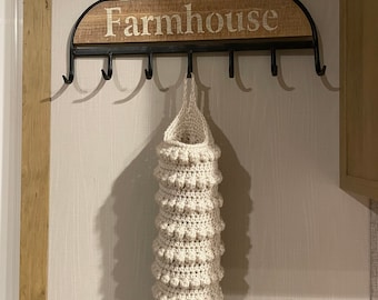 May include: A cream-colored crocheted bag with a textured bobble stitch pattern hanging from a black metal rack. The rack features a wooden panel with the word Farmhouse printed in white letters and includes eight black metal hooks.