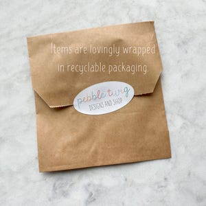 Pu&ograve; includere: Un pacco di carta marrone con il testo "Items are lovingly wrapped in recyclable packaging." e un adesivo che dice "pebble twig DESIGNS AND SHOP".