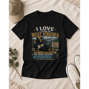 Puede incluir: Una camiseta negra con el gr&aacute;fico de un oso sentado en una mecedora y el texto I LOVE LIVING IN WEST VIRGINIA, WE DON'T DIAL 911 WE DIAL A NEIGHBOR, WHERE THE ROADS ARE WINDING, THE PEOPLE ARE KIND, AND THE ONLY TRAFFIC JAM IS BEHIND A SLOW TRACTOR.