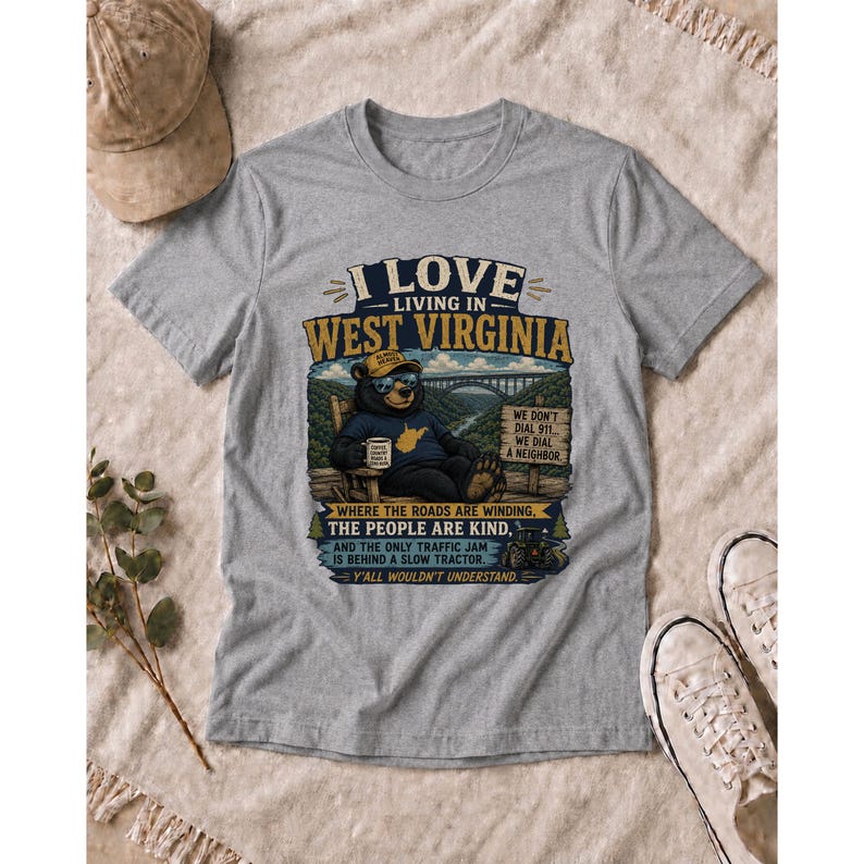 Puede incluir: Una camiseta gris jaspeado con un gr&aacute;fico de un oso con sombrero y gafas de sol, sentado en una silla con una taza. El texto dice I LOVE LIVING IN WEST VIRGINIA, WE DON'T DIAL 911, WE DIAL A NEIGHBOR y Y'ALL WOULDN'T UNDERSTAND.