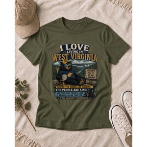 Puede incluir: Una camiseta verde oliva con el gr&aacute;fico de un oso negro con sombrero y gafas de sol, sentado en una silla con una taza. El texto dice I LOVE LIVING IN WEST VIRGINIA, WE DON'T DIAL 911, WE DIAL A NEIGHBOR y otras frases adicionales.