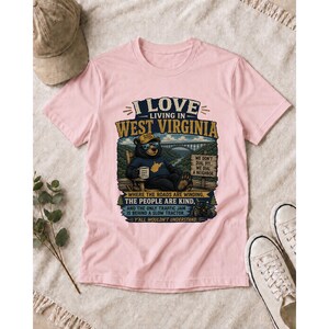 Puede incluir: Una camiseta rosa claro con el gr&aacute;fico de un oso con gafas de sol y sombrero, sentado en una silla con una taza. El texto dice I LOVE LIVING IN WEST VIRGINIA, WE DON'T DIAL 911, WE DIAL A NEIGHBOR, WHERE THE ROADS ARE KIND, Y'ALL WOULDN'T UNDERSTAND.