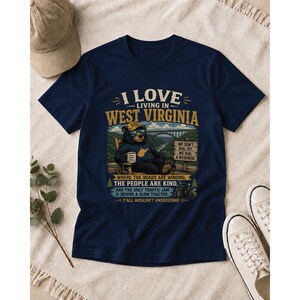 Puede incluir: Una camiseta azul marino con un gr&aacute;fico de un oso en una mecedora con gafas de sol y una gorra. El texto dice: I LOVE LIVING IN WEST VIRGINIA, WE DON'T DIAL 911, WE DIAL A NEIGHBOR, WHERE THE ROADS ARE WINDING, THE PEOPLE ARE KIND.