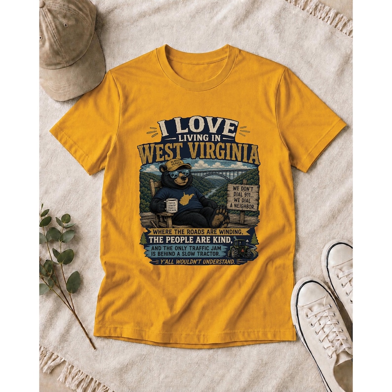 Puede incluir: Una camiseta de color amarillo mostaza con un gr&aacute;fico de un oso con gafas de sol y gorra, sentado en una silla. El texto dice: I LOVE LIVING IN WEST VIRGINIA, WE DON'T DIAL 911... WE DIAL A NEIGHBOR, WHERE THE ROADS ARE WINDING, THE PEOPLE ARE KIND.