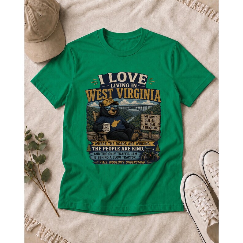 Puede incluir: Una camiseta verde de manga corta con un gr&aacute;fico de un oso sentado en una silla y el texto I LOVE LIVING IN WEST VIRGINIA, WE DON'T DIAL 911, WE DIAL A NEIGHBOR, WHERE THE ROADS ARE WINDING, THE PEOPLE ARE KIND, Y Y'ALL WOULDN'T UNDERSTAND.