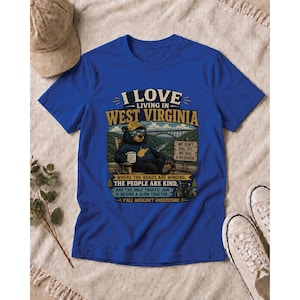 Puede incluir: Una camiseta azul real con el gr&aacute;fico de un oso con gafas de sol y sombrero, sentado en una silla con una taza. El texto dice I LOVE LIVING IN WEST VIRGINIA, WE DON'T DIAL 911, WE DIAL A NEIGHBOR, y Y'ALL WOULDN'T UNDERSTAND.
