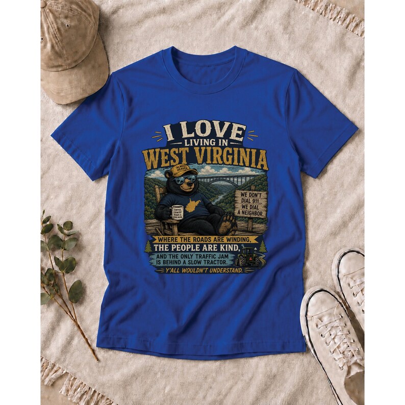 Puede incluir: Una camiseta azul real con el gr&aacute;fico de un oso con gafas de sol y sombrero, sentado en una silla con una taza. El texto dice I LOVE LIVING IN WEST VIRGINIA, WE DON'T DIAL 911, WE DIAL A NEIGHBOR, y Y'ALL WOULDN'T UNDERSTAND.