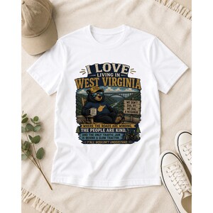 Puede incluir: Una camiseta blanca de manga corta con un gr&aacute;fico de un oso sentado en una silla y el texto I LOVE LIVING IN WEST VIRGINIA, WHERE THE ROADS ARE WINDING, THE PEOPLE ARE KIND, AND THE ONLY TRAFFIC JAM IS BEHIND A SLOW TRACTOR.