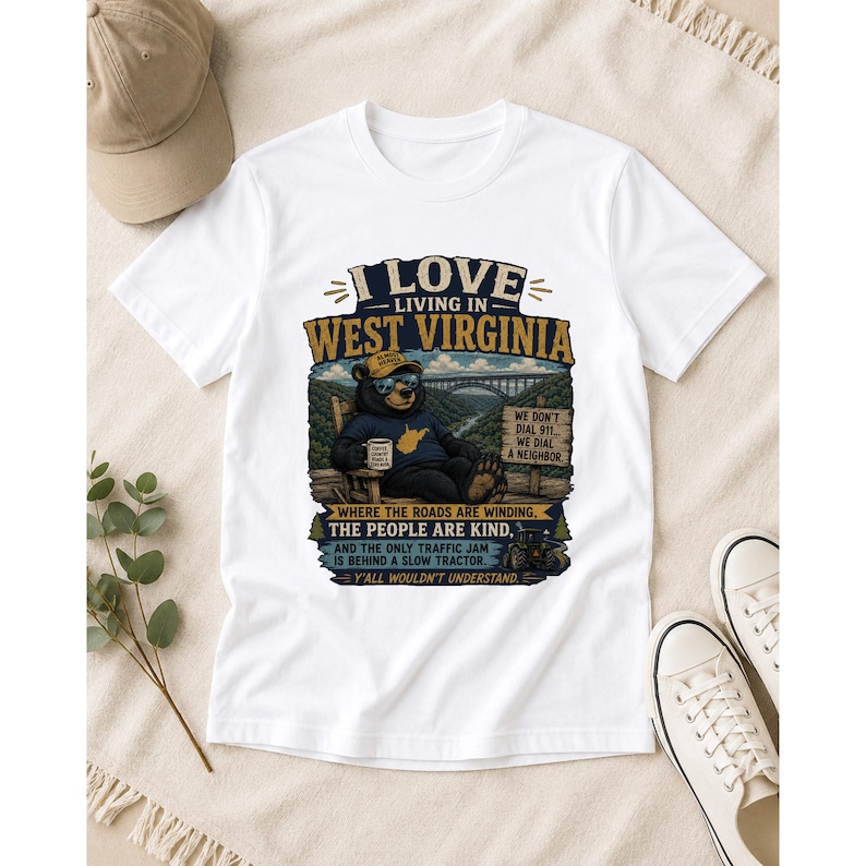 Puede incluir: Una camiseta blanca de manga corta con un gr&aacute;fico de un oso sentado en una silla y el texto I LOVE LIVING IN WEST VIRGINIA, WHERE THE ROADS ARE WINDING, THE PEOPLE ARE KIND, AND THE ONLY TRAFFIC JAM IS BEHIND A SLOW TRACTOR.