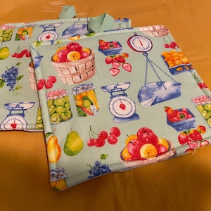 May include: Two square pot holders with a light green background. The fabric is patterned with illustrations of fruit, scales, and jars. The fruit includes apples, pears, cherries, and grapes. Each pot holder has a green loop for hanging.