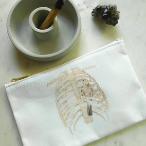 May include: A white canvas zipper pouch with a gold zipper, featuring a watercolor illustration of a rib cage. A concrete incense holder with a palo santo stick and a dark crystal are also visible.
