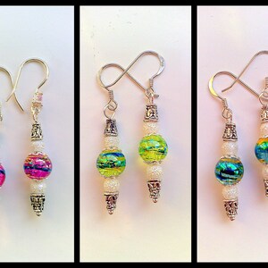 May include: Three pairs of dangle earrings with silver hooks and white beads. The earrings feature a large round glass bead with iridescent colors. The beads are pink, green, and blue.