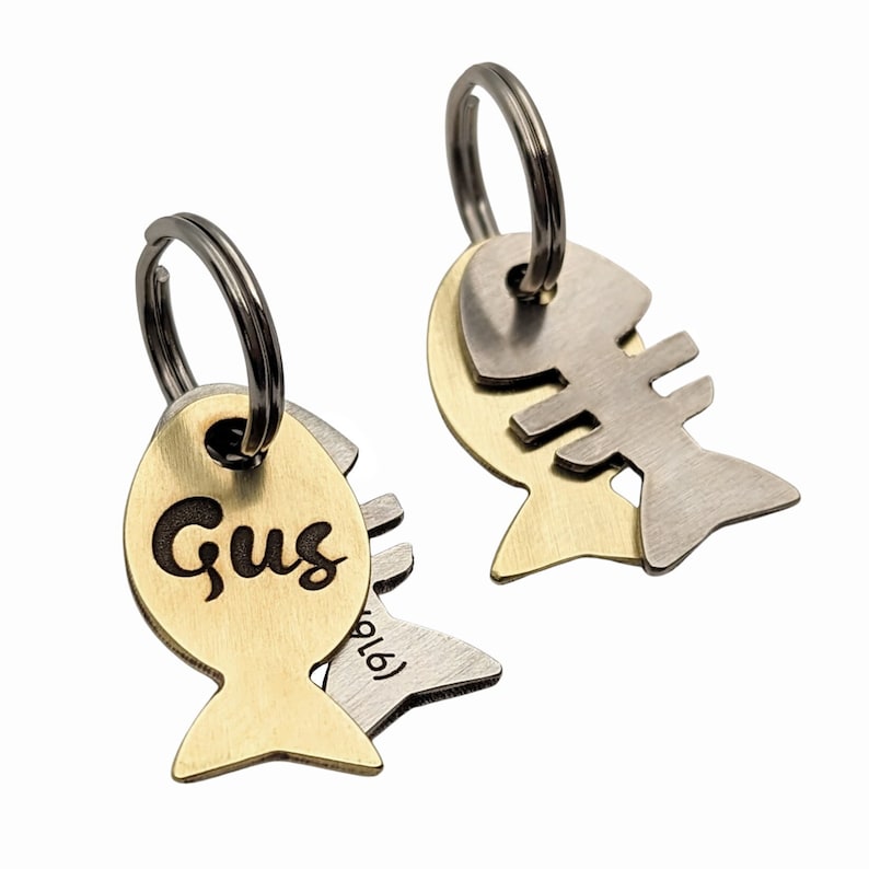 May include: Two gold and silver fish-shaped pet ID tags with key rings. The gold tag has the name "Gus" engraved on it. The silver tag has a fish skeleton design.