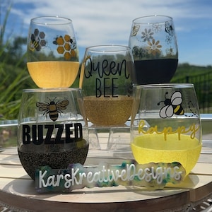 May include: Four wine glasses with bee-themed designs. The glasses are filled with a yellow liquid and have black glitter at the bottom. The designs include "Queen Bee", "Buzzed", "Happy", and a bee with honeycomb. A sign reads "Kars Kreative Designs".