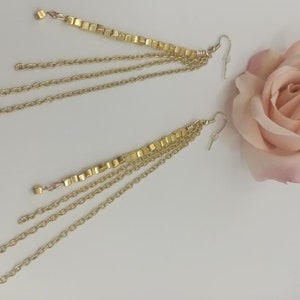 May include: A pair of gold earrings with multiple chains and a row of gold beads. The earrings are on a white surface with a pink rose.