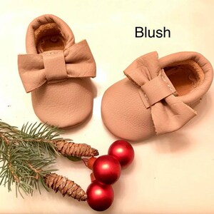 May include: A pair of blush-colored leather baby moccasins with a bow on the front. The moccasins are on a white surface with a pine branch and red ornaments.