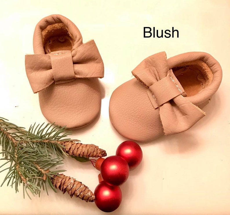 May include: A pair of blush-colored leather baby moccasins with a bow on the front. The moccasins are on a white surface with a pine branch and red ornaments.