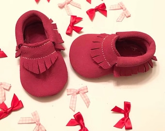 May include: A pair of red suede baby moccasins with fringe detailing and white stitching. Scattered around the shoes are small red and pink bows. The shoes are designed for infants and toddlers.