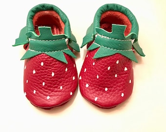 May include: A pair of baby shoes designed to resemble strawberries. The shoes are primarily red with white dot accents, green leaf-shaped details, and a green trim. The interior of the shoes is a light red color.