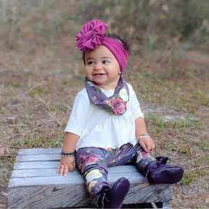 May include: A baby girl wearing a white shirt, floral leggings, and a purple headband with a large bow. She is also wearing a floral print scarf around her neck. The baby is sitting on a wooden platform.