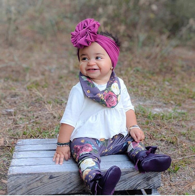 May include: A baby girl wearing a white shirt, floral leggings, and a purple headband with a large bow. She is also wearing a floral print scarf around her neck. The baby is sitting on a wooden platform.