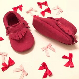 May include: Two pairs of red suede baby moccasins with fringe detailing. Scattered around the shoes are small red and pink bows. The moccasins are a soft, muted red color, and the bows add a playful touch.