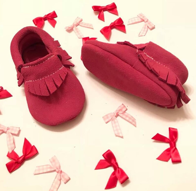 May include: Two pairs of red suede baby moccasins with fringe detailing. Scattered around the shoes are small red and pink bows. The moccasins are a soft, muted red color, and the bows add a playful touch.