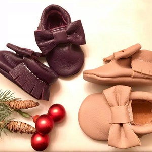 May include: Two pairs of leather baby moccasins with bows. One pair is purple and the other is light brown.