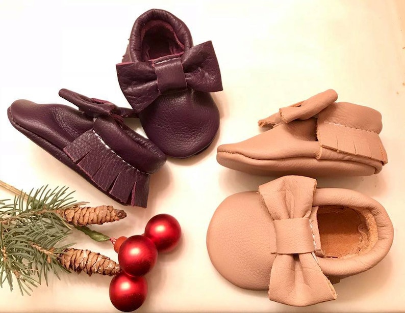 May include: Two pairs of leather baby moccasins with bows. One pair is purple and the other is light brown.
