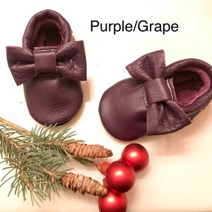 May include: A pair of purple leather baby moccasins with a bow on the top. The moccasins are on a white surface with a pine branch and red ornaments.