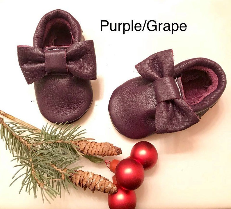May include: A pair of purple leather baby moccasins with a bow on the top. The moccasins are on a white surface with a pine branch and red ornaments.