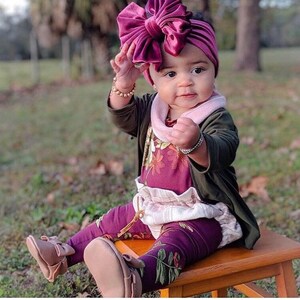 May include: A baby girl wearing a burgundy velvet headband with a large bow, a floral print top, a white ruffled skirt, and burgundy leggings with a floral print. She is wearing brown leather shoes and has a pink and white scarf around her neck.